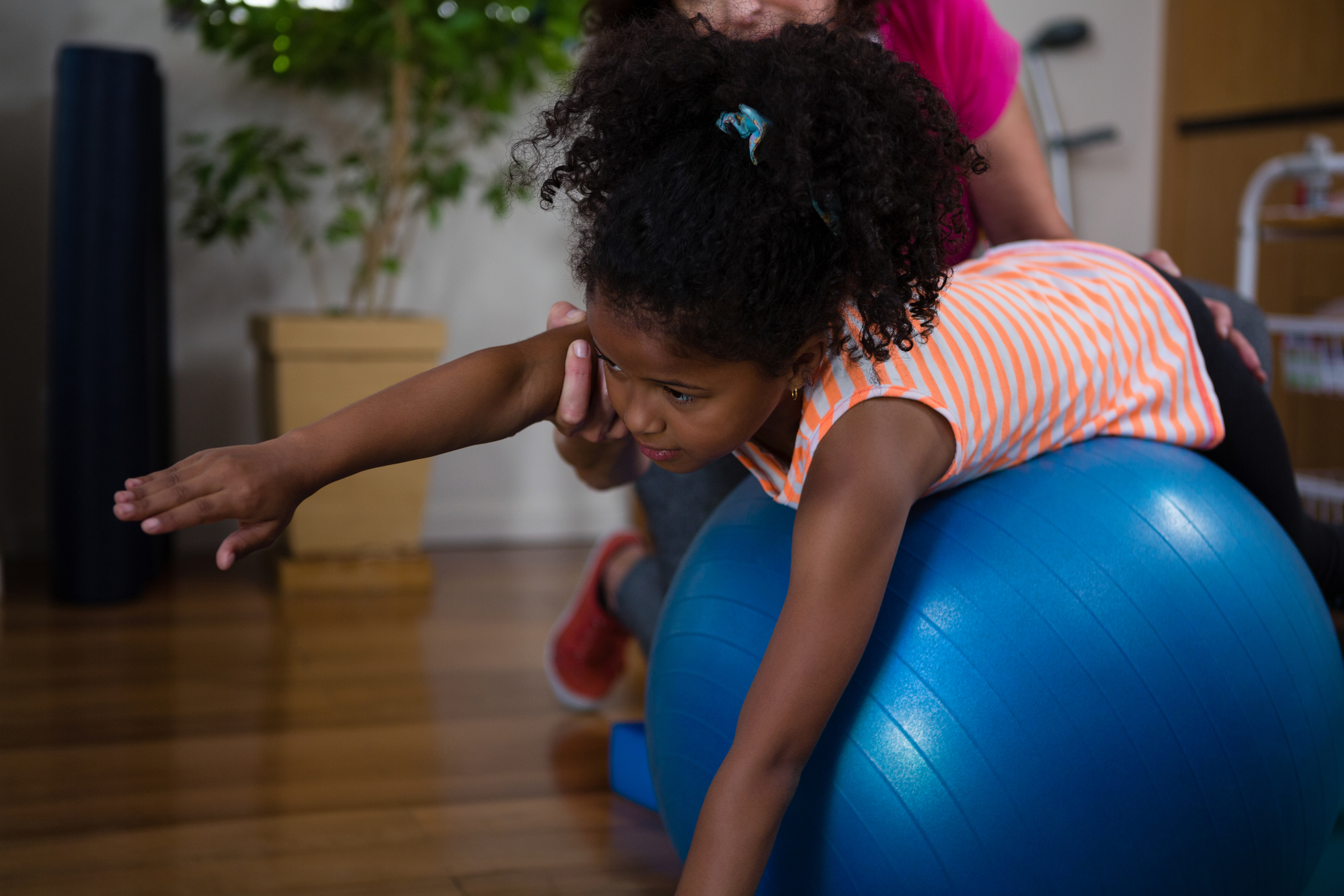 Female physiotherapist helping girl patient in performing exercise on fitness ball